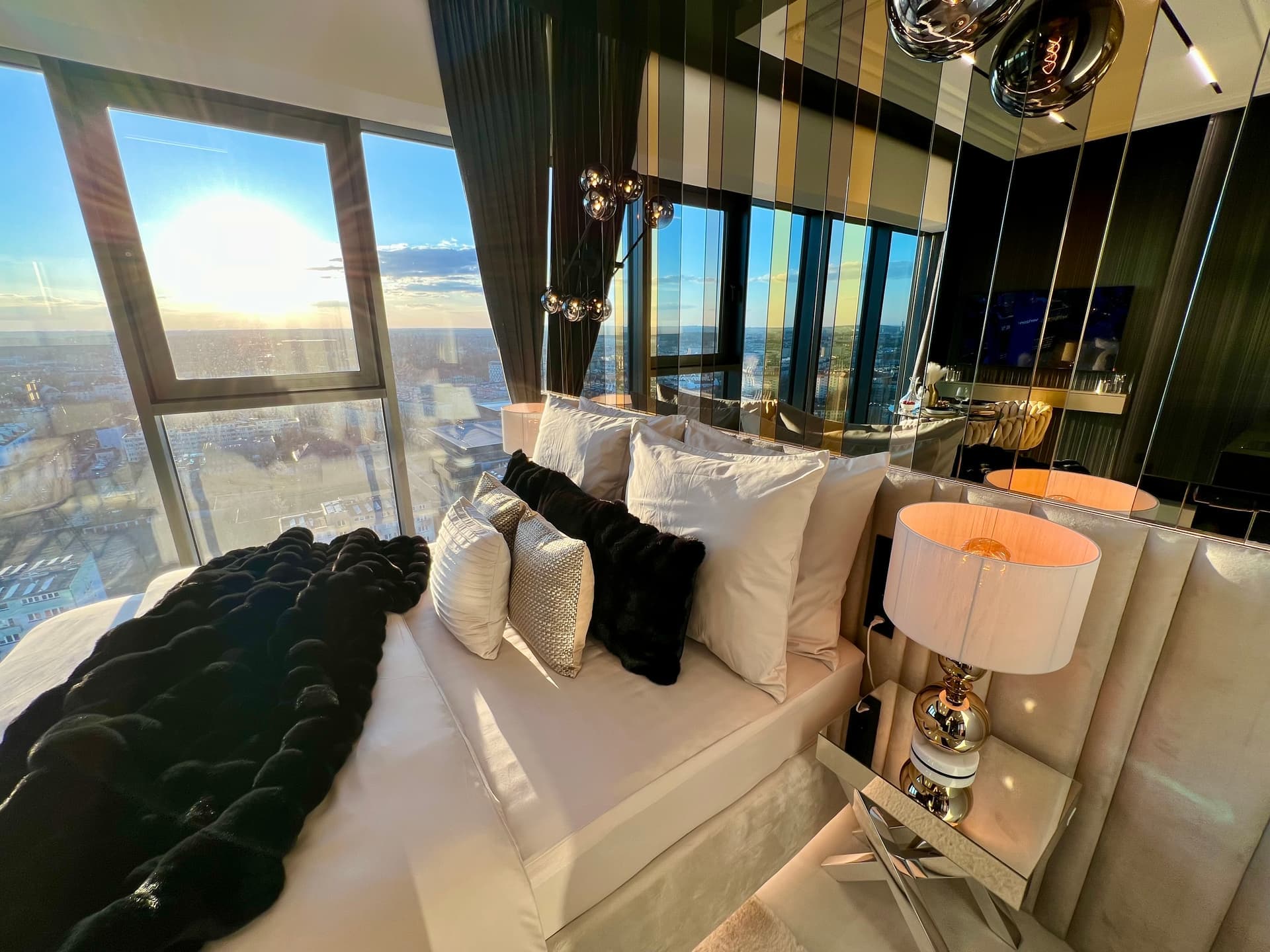 King-size bed with cushions and black throw, sunset through floor-to-ceiling windows