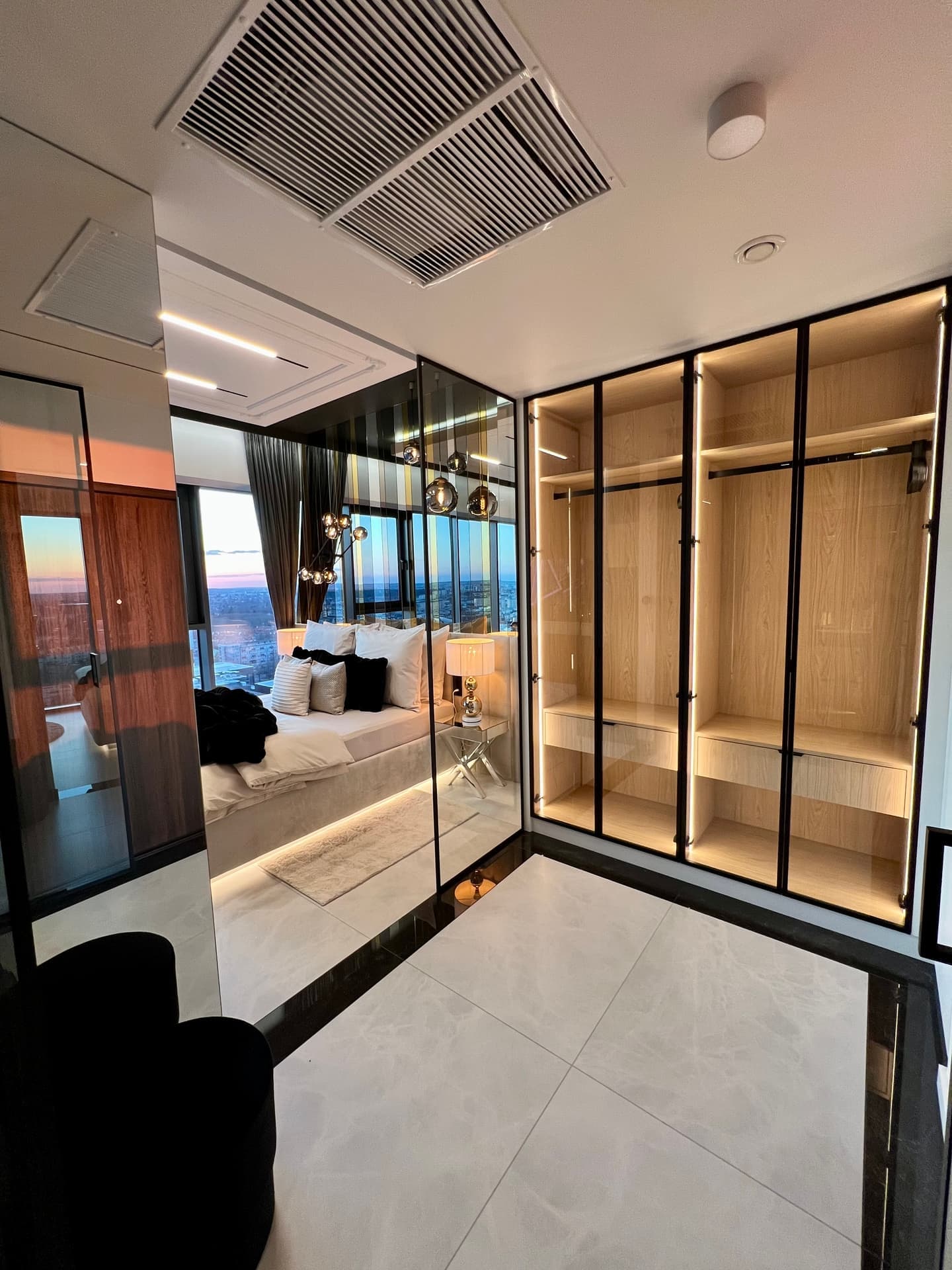 Walk-in closet with lit wardrobe, glass doors and mirror reflecting the bedroom